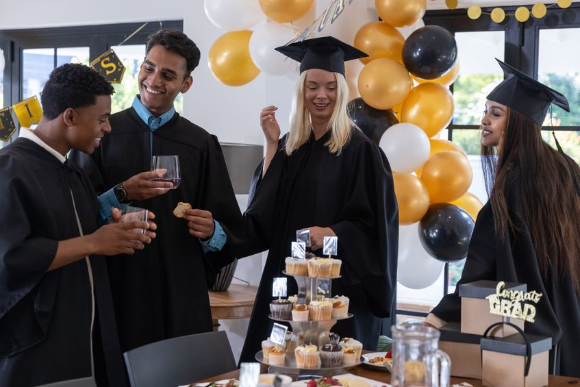 Friends in gowns gathering at table at graduation, sharing cupcakes and toasting wine.